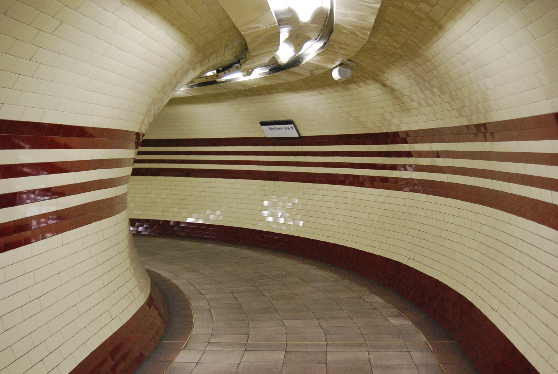 Hampstead tube station tunnel, empty, with cream and red tiles, curving around an corner.